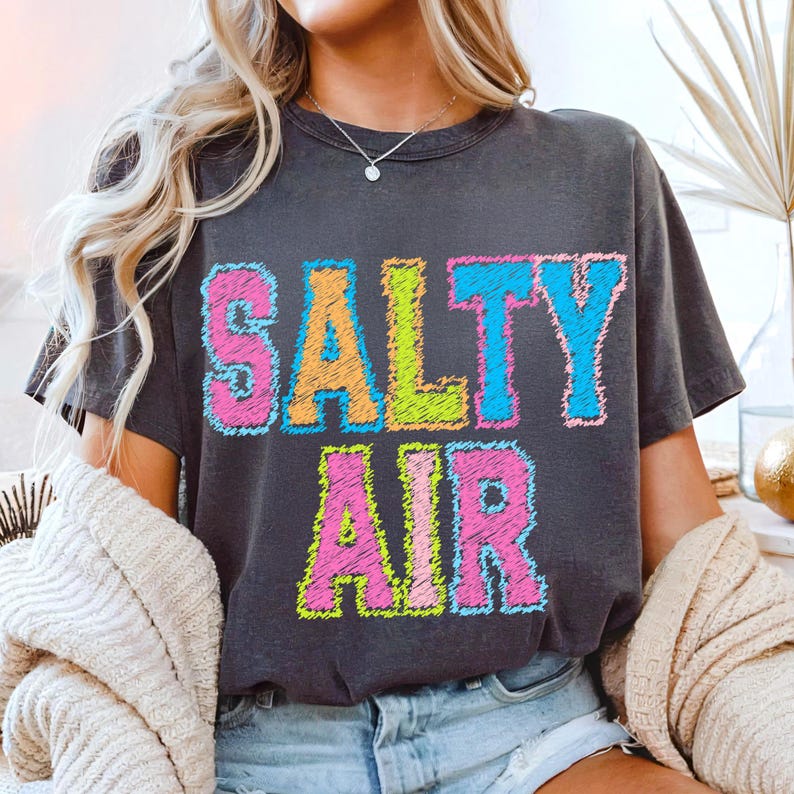 May include: Dark gray t-shirt with the words "SALTY AIR" in colorful, stitched-style lettering. The text features orange, green, pink, and blue colors. The shirt is a casual, comfortable style.