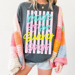 K&ouml;nnte beinhalten: Dunkelgraues Sweatshirt mit dem Text "Made for Sunny Days" in T&uuml;rkis, Rosa und Gelb. Das Design umfasst eine rosa Schleife und vertikale rosa Streifen. Dar&uuml;ber wird eine bunte Patchwork-Strickjacke getragen.