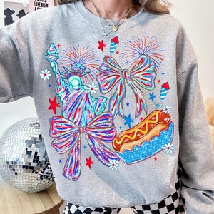 May include: A gray sweatshirt featuring a colorful graphic of the Statue of Liberty, bows, fireworks, and a hot dog. The design incorporates red, white, and blue elements, with stars and flowers. The sweatshirt has a crew neck.