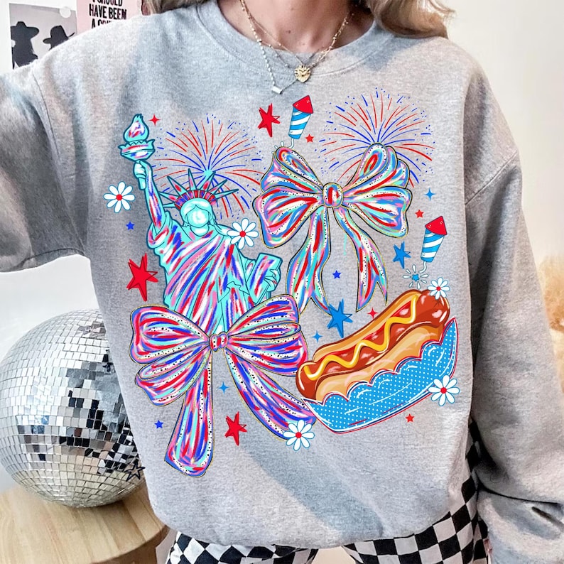 May include: A gray sweatshirt featuring a colorful graphic of the Statue of Liberty, bows, fireworks, and a hot dog. The design incorporates red, white, and blue elements, with stars and flowers. The sweatshirt has a crew neck.