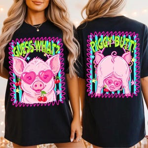 Op de afbeelding: Zwart T-shirt met een roze varken met een hartvormige zonnebril en kersen. De voorkant van het shirt heeft de tekst "GUESS WHAT?" in groen, en de achterkant "PIGGY BUTT!" ook in groen.