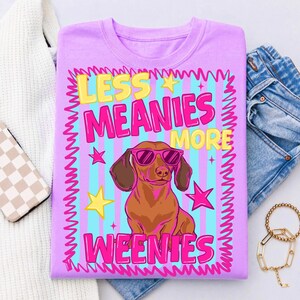 May include: A lilac t-shirt with a cartoon dachshund wearing pink sunglasses. The text "LESS MEANIES MORE WEENIES" is in yellow and pink, with stars and a striped background. The design is framed.
