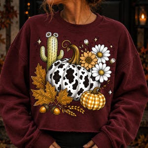 Puede incluir: Una sudadera de manga larga color granate con un estampado gr&aacute;fico de una calabaza con manchas de vaca, una calabaza de cuadros amarillos, un cactus verde, margaritas blancas, un girasol amarillo, hojas de oto&ntilde;o marrones y dos bellotas.