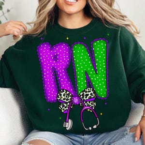 May include: Dark green sweatshirt with a large graphic featuring the letters "RN" in purple and green polka dots. A leopard print bow and a purple stethoscope are below the letters. The design includes decorative purple trim.