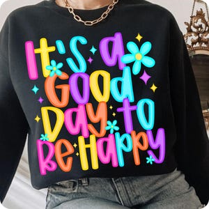 K&ouml;nnte beinhalten: Schwarzer Pullover mit dem Text "It's a good day to be happy" in bunten, umrandeten Buchstaben. Der Text ist mit kleinen Blumen und Sternen verziert.