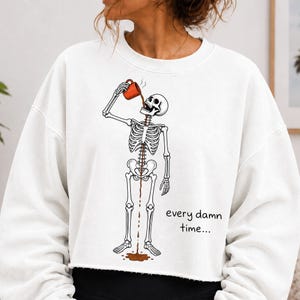 White folded t-shirt with a black line-art skeleton pouring coffee from a red mug into its open mouth, coffee dripping down to a puddle at its feet, and the words every damn time in black handwriting. Funny coffee lover sublimation design flat lay with a straw hat.