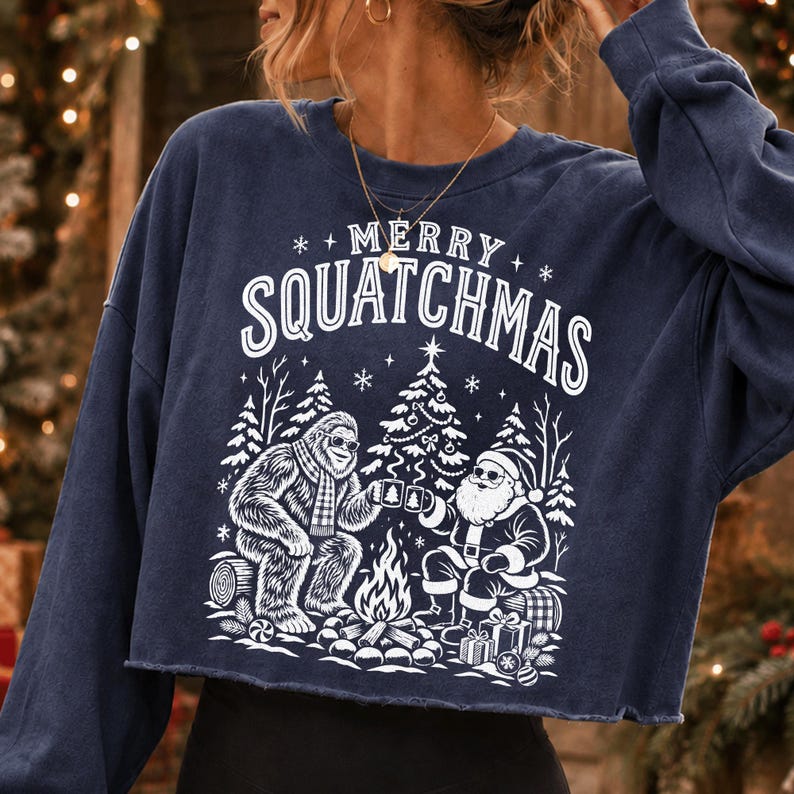 K&ouml;nnte beinhalten: Ein marineblaues Langarm-Sweatshirt mit offenem Saum und wei&szlig;em Grafikdruck. Das Motiv zeigt einen Bigfoot und den Weihnachtsmann, die an einem Lagerfeuer sitzen, mit dem Text MERRY SQUATCHMAS dar&uuml;ber, umgeben von Tannenb&auml;umen und Schneeflocken.