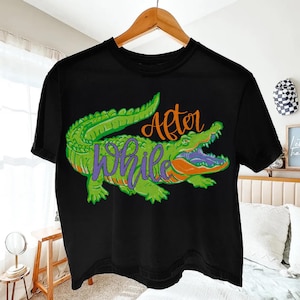 Pu&ograve; includere: T-shirt nero con un design di alligatore verde, dettagli arancioni e testo viola che recita "After While". L'alligatore ha la bocca aperta. Il design &egrave; dettagliato e vibrante, creando una grafica audace.