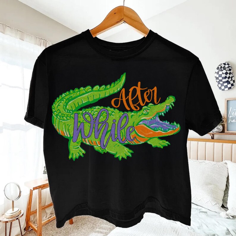 Pu&ograve; includere: T-shirt nero con un design di alligatore verde, dettagli arancioni e testo viola che recita "After While". L'alligatore ha la bocca aperta. Il design &egrave; dettagliato e vibrante, creando una grafica audace.