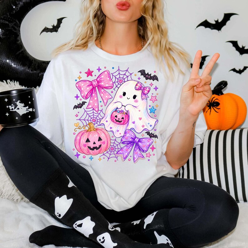 Peut inclure: Un t-shirt blanc avec une impression graphique color&eacute;e repr&eacute;sentant un fant&ocirc;me mignon tenant un petit seau citrouille, entour&eacute; de n&oelig;uds roses et violets, de toiles d'araign&eacute;e, d'&eacute;toiles et de chauves-souris, avec une grande citrouille rose.
