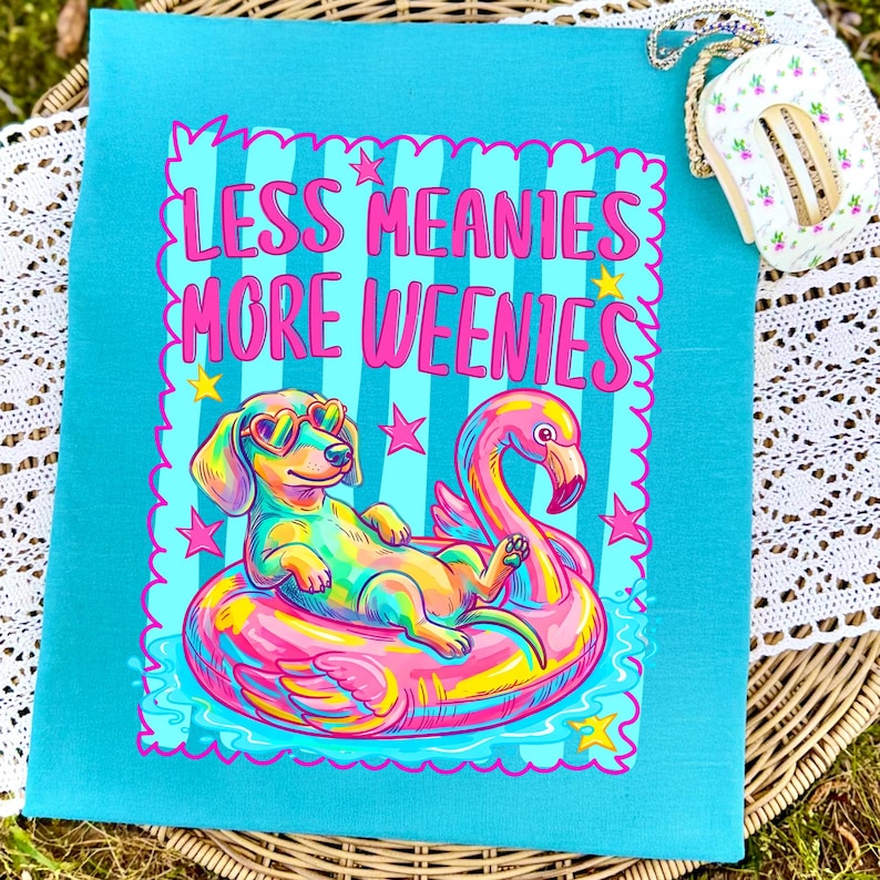 May include: A bright turquoise t-shirt featuring a colorful graphic of a dachshund wearing heart-shaped sunglasses while floating on a pink flamingo pool float. The text "LESS MEANIES MORE WEENIES" is printed in pink above the dog, surrounded by small yellow and pink stars.