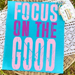 May include: A turquoise canvas with the words "FOCUS ON THE GOOD" printed in pink and purple. The text has a distressed, textured appearance. A small, floral-patterned pouch with a gold chain is visible in the upper right corner.