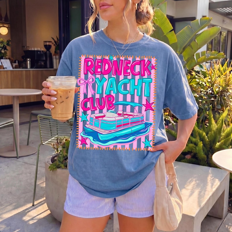 May include: Blue t-shirt with the text "Redneck Yacht Club" and a graphic of a pontoon boat. The design features pink, teal, and white colors. The shirt is worn with striped shorts.