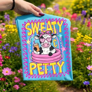 May include: A light blue t-shirt with the words "SWEATY PETTY" in yellow and pink lettering. The shirt features a cartoon cow wearing heart-shaped sunglasses and a pink headband, floating in a pink inner tube and holding a drink.