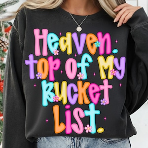 K&ouml;nnte beinhalten: Ein schwarzes Sweatshirt mit dem Text "Heaven. top of my bucket list." in bunten Buchstaben. Der Text ist mit kleinen Blumen verziert. Das Sweatshirt hat einen l&auml;ssigen Stil.