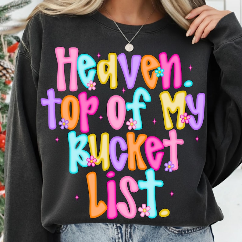 K&ouml;nnte beinhalten: Ein schwarzes Sweatshirt mit dem Text "Heaven. top of my bucket list." in bunten Buchstaben. Der Text ist mit kleinen Blumen verziert. Das Sweatshirt hat einen l&auml;ssigen Stil.