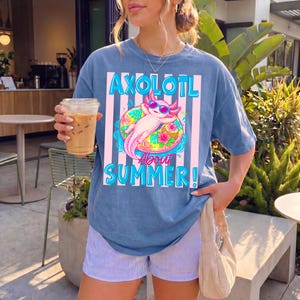 Peut inclure: Un t-shirt bleu avec le texte "AXOLOTL ABOUT SUMMER!" et un graphisme d'axolotl rose portant des lunettes de soleil sur un bou&eacute;e. Le t-shirt est port&eacute; avec un short &agrave; rayures.