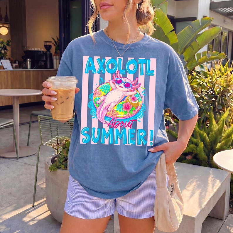 Peut inclure: Un t-shirt bleu avec le texte "AXOLOTL ABOUT SUMMER!" et un graphisme d'axolotl rose portant des lunettes de soleil sur un bou&eacute;e. Le t-shirt est port&eacute; avec un short &agrave; rayures.