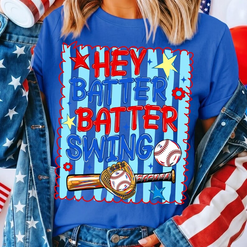 K&ouml;nnte beinhalten: Blaues T-Shirt mit dem Text "HEY BATTER BATTER SWING" in Rot und Blau. Das Design umfasst einen Baseball, einen Schl&auml;ger und einen Handschuh. Das Shirt hat einen gestreiften Hintergrund mit Sternen und Blumen.
