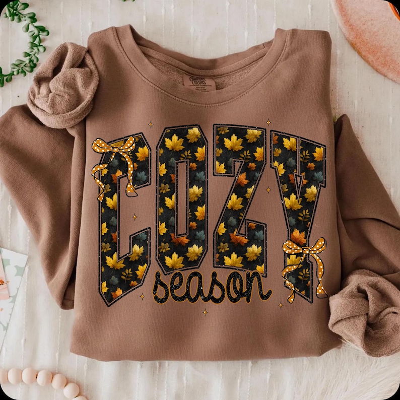 May include: A brown crewneck sweatshirt featuring the text COZY in large block letters filled with a pattern of yellow and orange autumn leaves. The word season is written below in black script. Two yellow polka dot bows accent the letters.