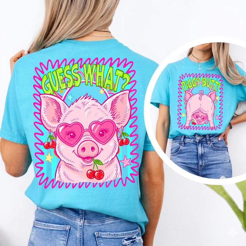 Op de afbeelding: Lichtblauw T-shirt met een roze varken met een hartvormige zonnebril en kersen. De tekst "GUESS WHAT?" staat boven het varken in een groene en roze golfvormige rand. Een kleinere afbeelding toont een vergelijkbaar shirt met de tekst "PIGGY BUTT!"