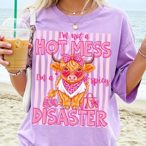 K&ouml;nnte beinhalten: Ein lila T-Shirt mit einer Grafik einer Highland-Kuh mit herzf&ouml;rmiger Sonnenbrille und Schleife. Der Text auf dem Shirt lautet: "I'm not a HOT MESS, I'm a spicy DISASTER."