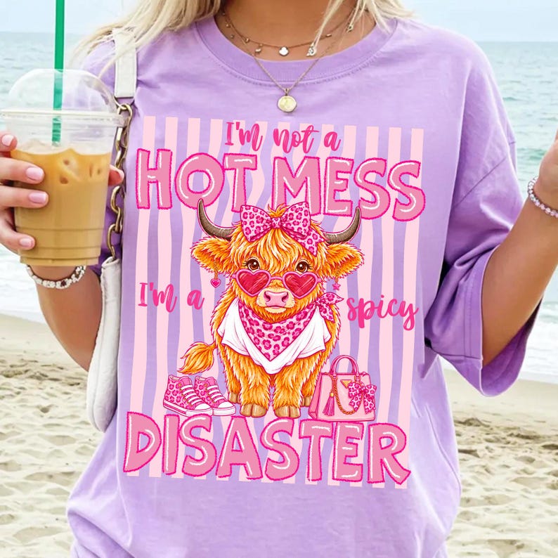 K&ouml;nnte beinhalten: Ein lila T-Shirt mit einer Grafik einer Highland-Kuh mit herzf&ouml;rmiger Sonnenbrille und Schleife. Der Text auf dem Shirt lautet: "I'm not a HOT MESS, I'm a spicy DISASTER."