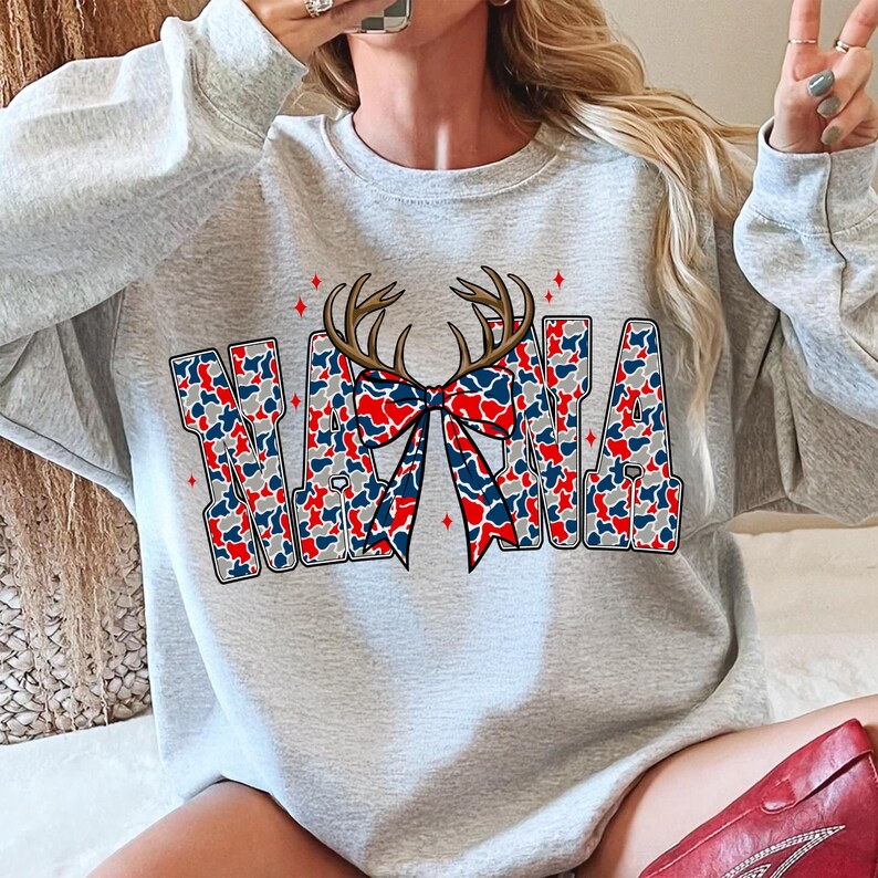 Peut inclure: Un sweat-shirt gris clair avec le mot "NANA" dans un motif camouflage rouge, blanc et bleu. Les lettres sont orn&eacute;es d'un motif de bois de cerf marron et d'un n&oelig;ud rouge, blanc et bleu.