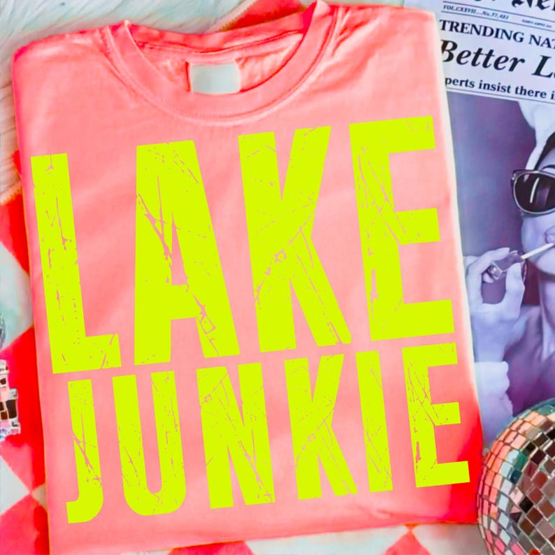 May include: A folded coral-colored t-shirt with the words "LAKE JUNKIE" printed in large, neon yellow letters. The shirt is on a patterned surface with a newspaper in the background.