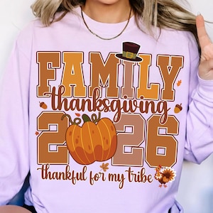 Pode incluir: Uma camisola de manga comprida em roxo claro com um gr&aacute;fico que inclui o texto "FAMILY thanksgiving 2026" e "thankful for my tribe". O design apresenta uma ab&oacute;bora castanha, um chap&eacute;u de peregrino, bolotas, folhas e um peru.