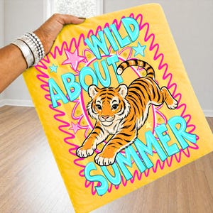 K&ouml;nnte beinhalten: Gelbe Decke mit einer Cartoon-Tiger-Illustration. Auf der Decke stehen die Worte "WILD ABOUT SUMMER" in T&uuml;rkis und Pink. Der Tiger ist orange und schwarz.