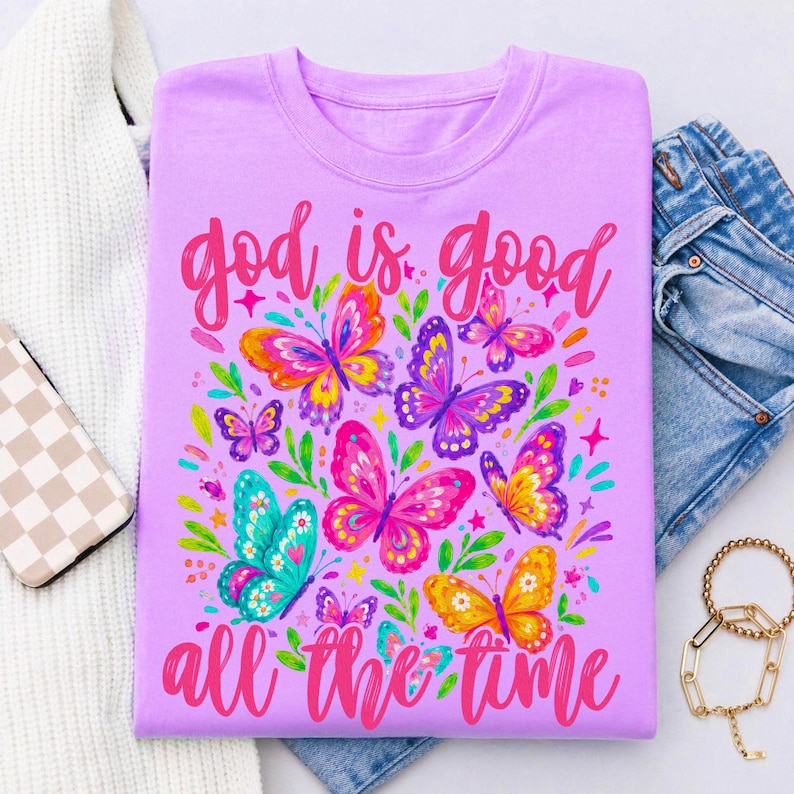 May include: A light purple t-shirt featuring a colorful graphic of multiple butterflies in shades of pink, yellow, blue, and purple, surrounded by green leaves and small stars, with the text "god is good all the time" in a pink script font.