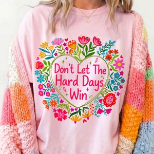 K&ouml;nnte beinhalten: Hellrosa T-Shirt mit floralem Herzdesign und dem Text "Don't Let The Hard Days Win". Das Herz ist von bunten Blumen und gr&uuml;nen Bl&auml;ttern umgeben. Die &Auml;rmel eines mehrfarbigen Pullovers sind sichtbar.