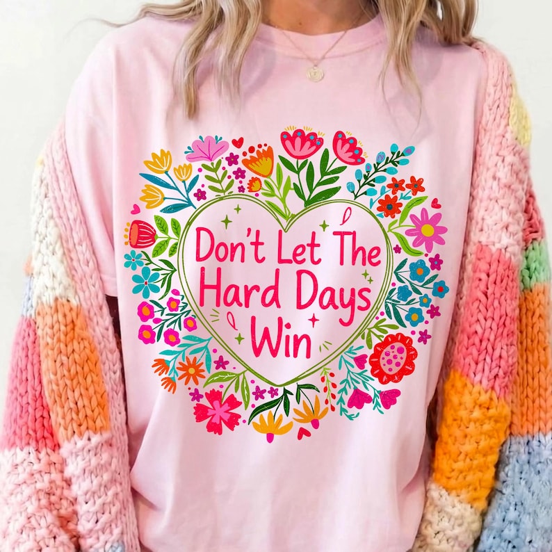 K&ouml;nnte beinhalten: Hellrosa T-Shirt mit floralem Herzdesign und dem Text "Don't Let The Hard Days Win". Das Herz ist von bunten Blumen und gr&uuml;nen Bl&auml;ttern umgeben. Die &Auml;rmel eines mehrfarbigen Pullovers sind sichtbar.