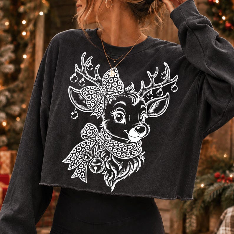 May include: A charcoal grey long-sleeve cropped sweatshirt featuring a white graphic print of a reindeer. The reindeer has a leopard print bow on its head and a leopard print scarf around its neck with two jingle bells attached to the scarf.