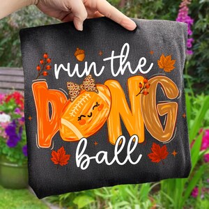 Puede incluir: Una camiseta gris oscuro con el texto blanco "run the" y "ball", con un gran gr&aacute;fico de bal&oacute;n de f&uacute;tbol americano naranja que sustituye a la "O" de "DONG". El bal&oacute;n tiene un lazo con estampado de leopardo, una cara sonriente y hojas oto&ntilde;ales.
