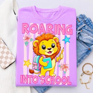 May include: A light purple t-shirt featuring a cartoon lion holding a pencil and a book, with a backpack on its back. The graphic includes pink and white vertical stripes, flowers, stars, and the text ROARING INTO SCHOOL in pink lettering.