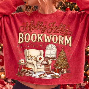 Op de afbeelding: Een rood sweatshirt met ronde hals en een grafische print met de tekst Holly Jolly BOOKWORM. De illustratie toont een gezellige fauteuil met een slapende kat, een open boek, een kerstboom, cadeautjes en een raam met een besneeuwd landschap.