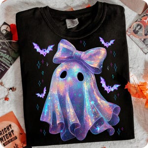 Może przedstawiać: Czarny t-shirt z fantazyjnym wzorem ducha. Duch jest ozdobiony dużą, opalizującą kokardą i otoczony fioletowymi nietoperzami i gwiazdami. Zabawny i świąteczny design na Halloween.