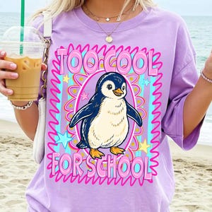 K&ouml;nnte beinhalten: Helllila T-Shirt mit Pinguin-Grafik. Das Design enth&auml;lt den Text "TOO COOL FOR SCHOOL" in Rosa, der einen Cartoon-Pinguin umgibt. Das T-Shirt hat kurze &Auml;rmel und eine lockere Passform.