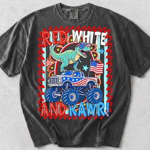 May include: Dark gray t-shirt featuring a graphic of a dinosaur wearing sunglasses and an American flag on a monster truck. The text reads "RED WHITE AND RAWR!" in red and white lettering. The design is centered on the shirt.