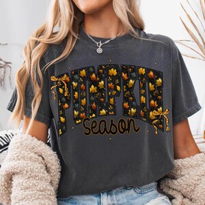 May include: A charcoal gray short-sleeve t-shirt featuring the text PUMPKIN SEASON in a bold, block-letter font. The letters are filled with a pattern of orange and yellow autumn leaves and are accented by two small, decorative yellow bows.