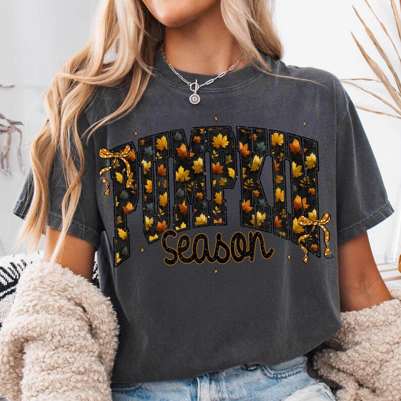 May include: A charcoal gray short-sleeve t-shirt featuring the text PUMPKIN SEASON in a bold, block-letter font. The letters are filled with a pattern of orange and yellow autumn leaves and are accented by two small, decorative yellow bows.