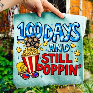 Puede incluir: Una toalla azul claro con el texto "100 DAYS AND STILL POPPIN'" en azul y rojo. El dise&ntilde;o incluye una cubeta de palomitas de ma&iacute;z, una bobina de pel&iacute;cula, gafas 3D y granos de palomitas.