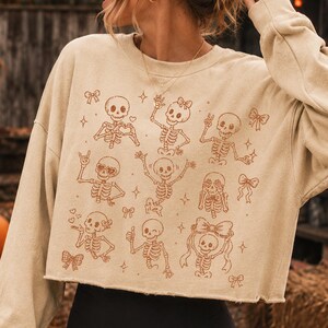 Op de afbeelding: Een beige cropped sweatshirt met een bruine grafische print van meerdere skeletten in verschillende houdingen, waaronder zwaaiend, hartjes vormend en een duim omhoog, afgewisseld met kleine illustraties van strikjes en sterren.