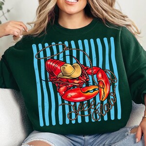 Op de afbeelding: Een donkergroene sweatshirt met een rode kreeft met een gele cowboyhoed, een touw en de woorden "Yee Claw" in een sierlijk lettertype. De kreeft staat tegen een blauw gestreepte achtergrond.