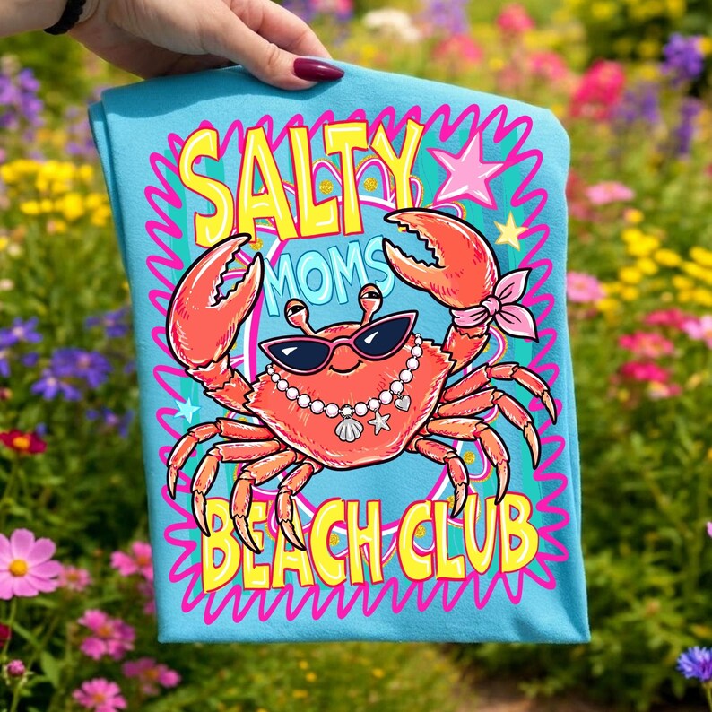 May include: A light blue t-shirt with a cartoon crab design. The crab wears sunglasses and a pearl necklace, with orange claws. The text "SALTY MOMS BEACH CLUB" is in yellow and pink. The shirt is held up by a hand, with a background of flowers.