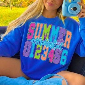 May include: A blue sweatshirt with the word "SUMMER" in large, colorful block letters. Below it, the word "Alphabet" is written in a cursive font, and the numbers 0 through 6 are displayed. The sweatshirt is a vibrant, eye-catching design.
