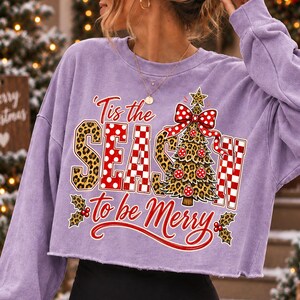 Op de afbeelding: Een lichtpaars sweatshirt met lange mouwen en een rafelige zoom. De afbeelding bevat de tekst 'Tis the SEASON to be Merry' in rood en wit, met een kerstboom met luipaardprint, een rode strik met stippen en hulstblaadjes als accenten.