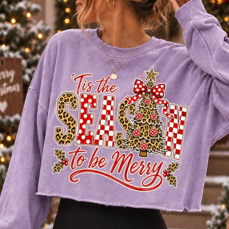 Op de afbeelding: Een lichtpaars sweatshirt met lange mouwen en een rafelige zoom. De afbeelding bevat de tekst 'Tis the SEASON to be Merry' in rood en wit, met een kerstboom met luipaardprint, een rode strik met stippen en hulstblaadjes als accenten.